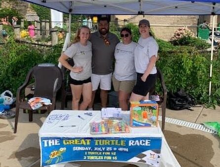 A group of 4 volunteers are at the Turtle Splash Water Park at a table advertising the great turtle race. There are toys and prizes on the table.