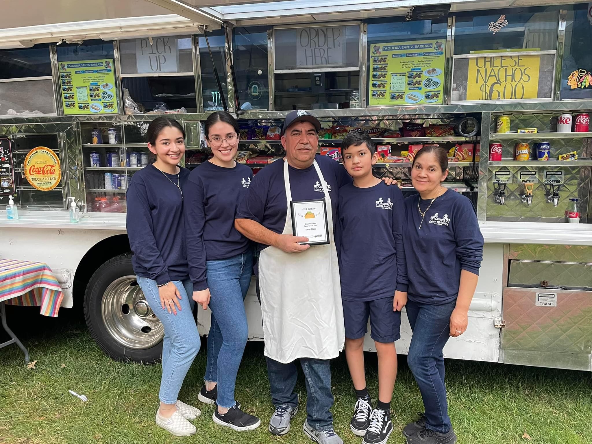 Five people are standing in front of a taco food truck. The owner of the food truck is holding a plaque declaring them the winners of the Taco Wars held at Reed Keppler Park.