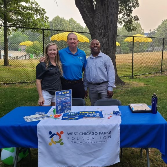 Three people are standing at a table in the park. There is a table cloth that says West Chicago Parks Foundation, and there are flyers on the table advertising the Turtle Race.