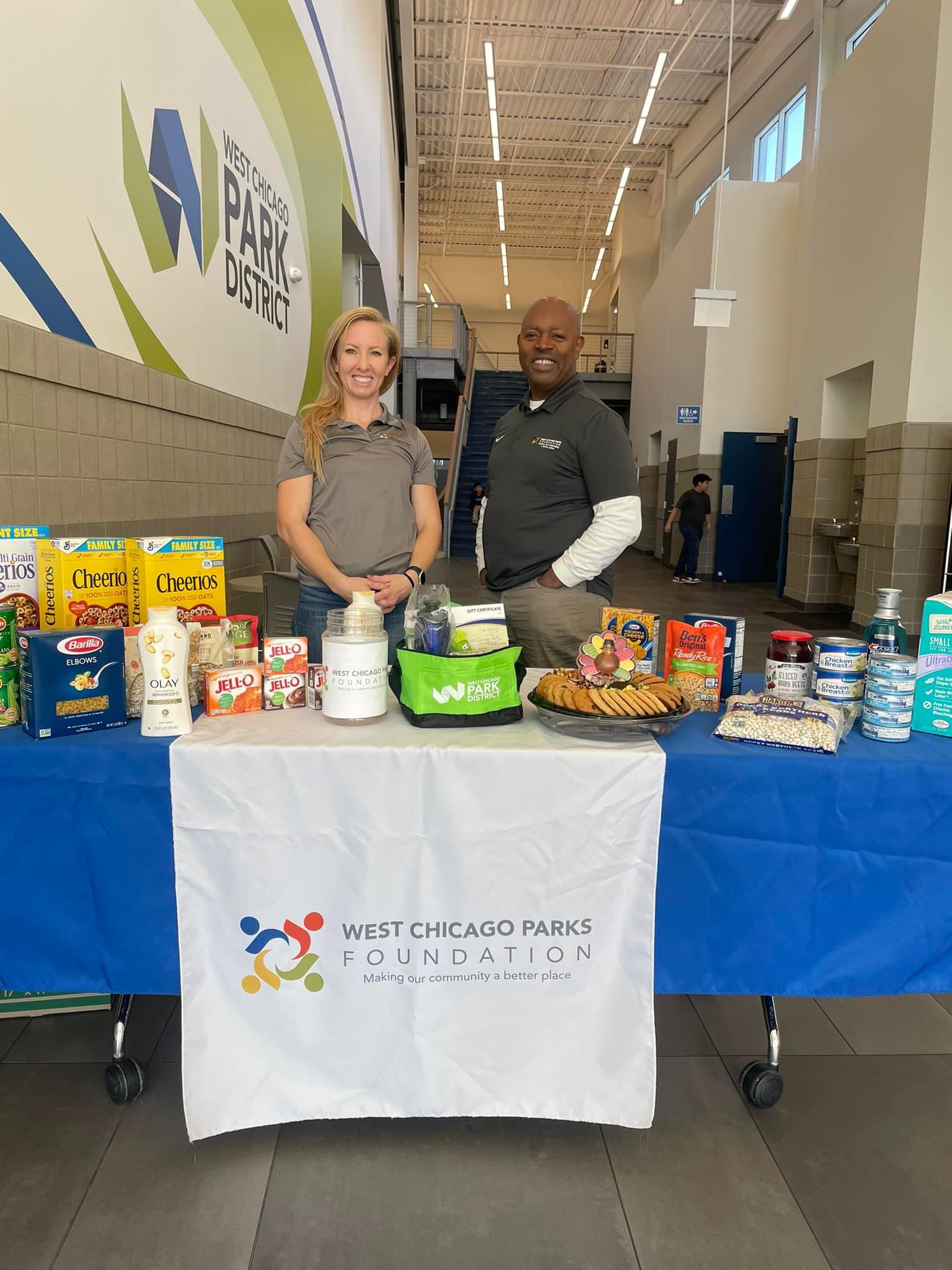 Two volunteers are standing at a table for the West Chicago Parks Foundation collecting food items for a food drive benefitting the local food pantry.