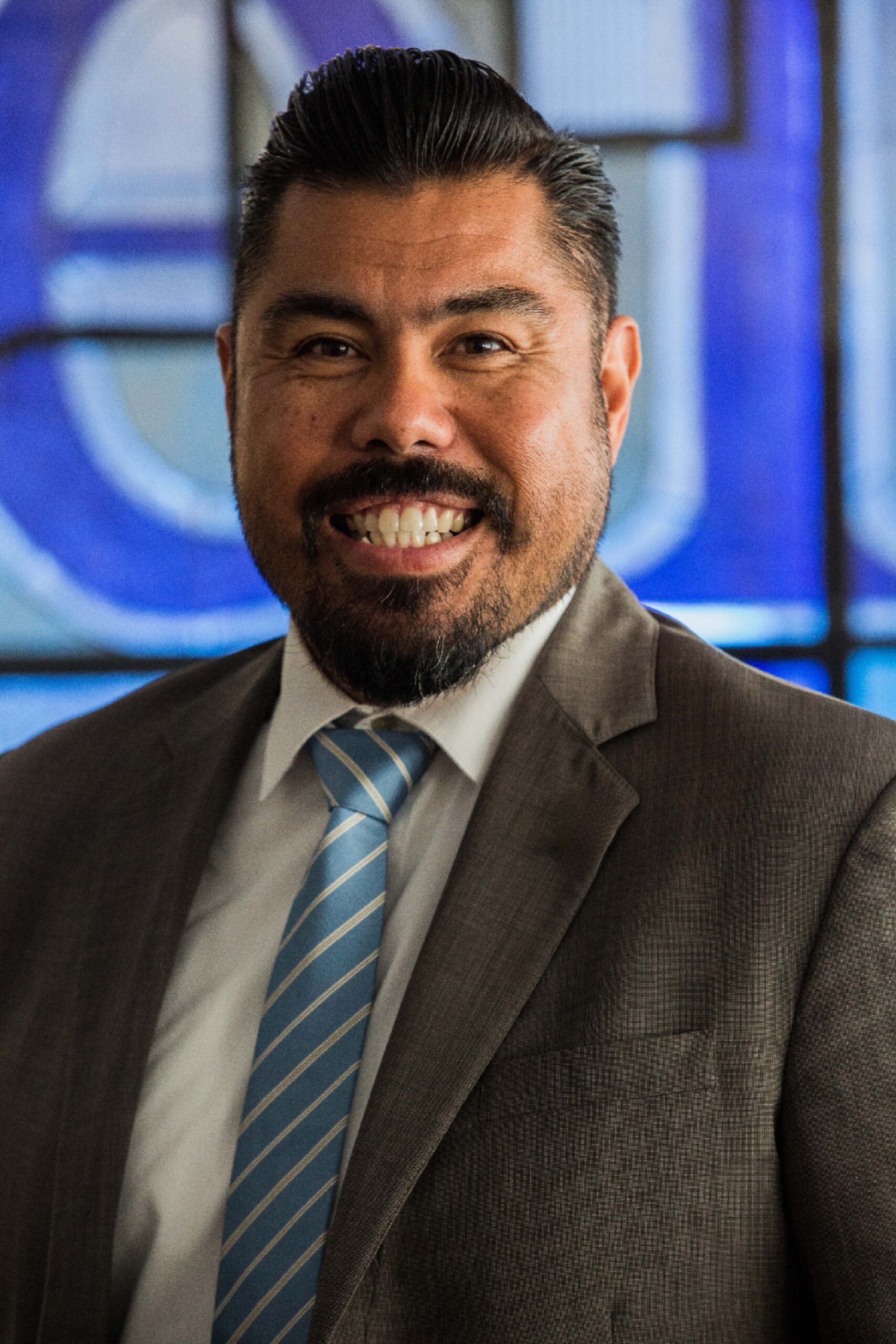 A headshot of Fil Guipoco. He is smiling and wearing a suit and tie against a blue background.