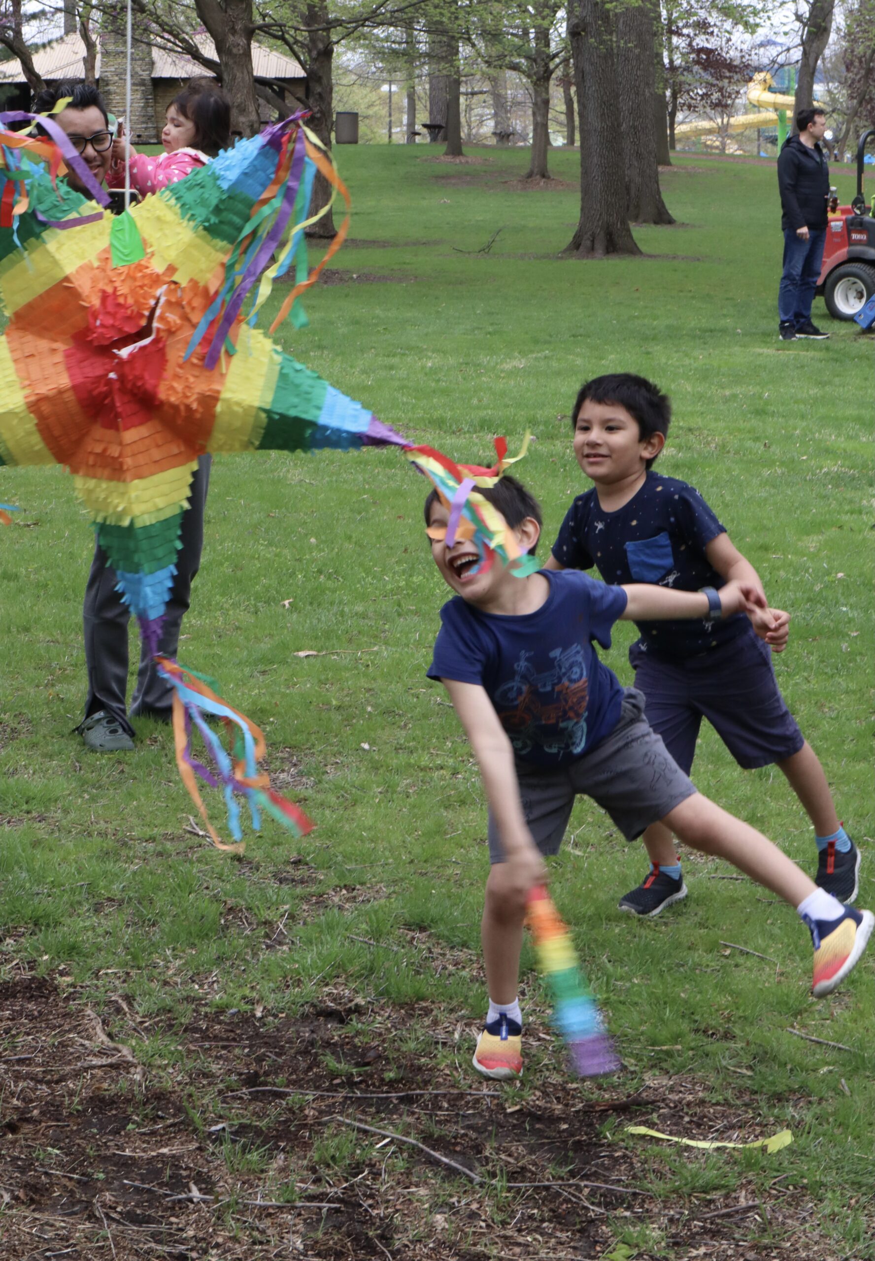 Two young children are smiling, trying to swing at a pinata. A father and his daughter watch as the children are having fun.