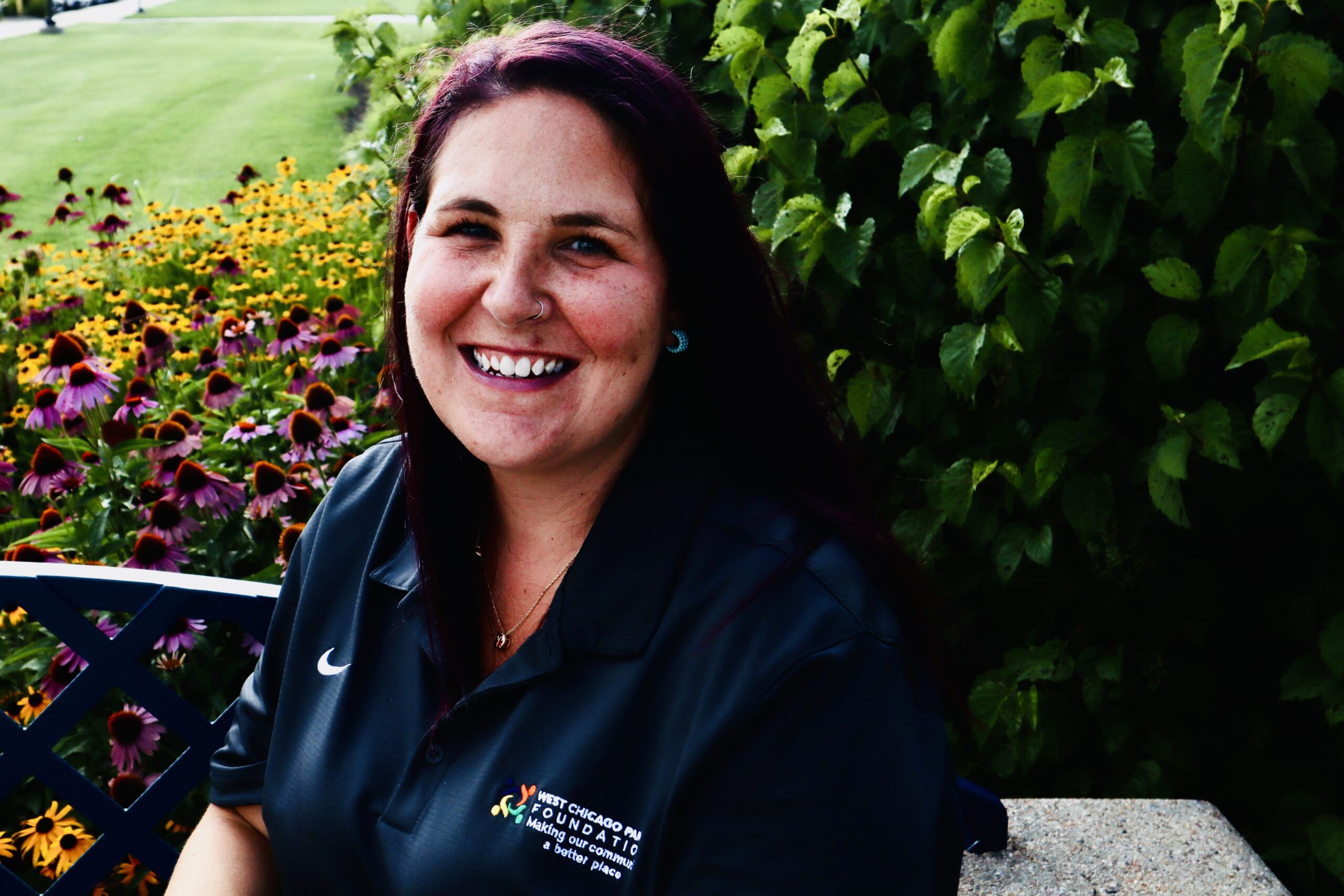 A headshot of Board Memeber, Sara Witteck. She is wearing a Parks Foundation shirt, smiling seated outside on a sunny day with foliage in the background