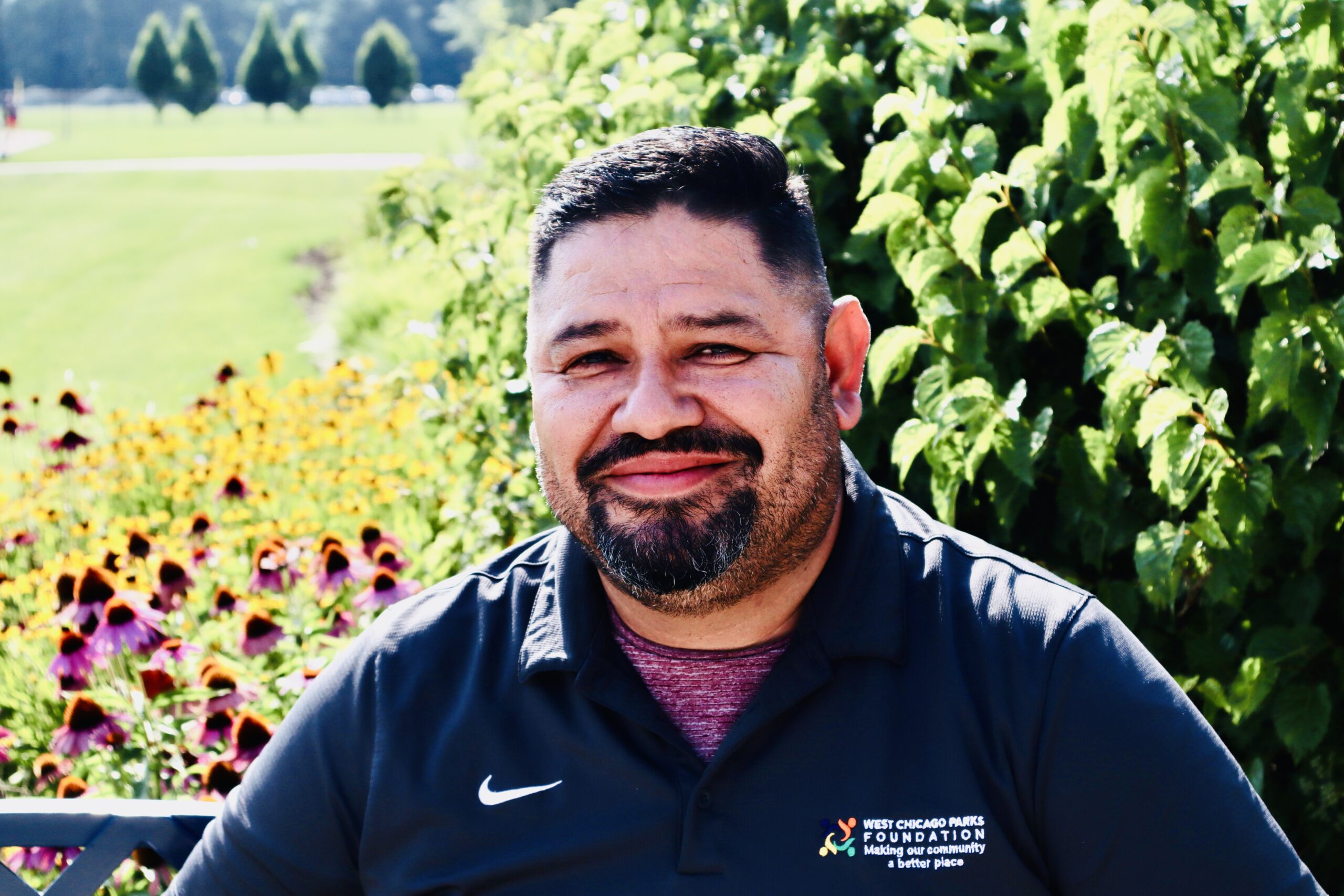 A headshot of Board Treasurer, Omar Espinosa. He is smiling seated outside on a sunny day with foliage in the background