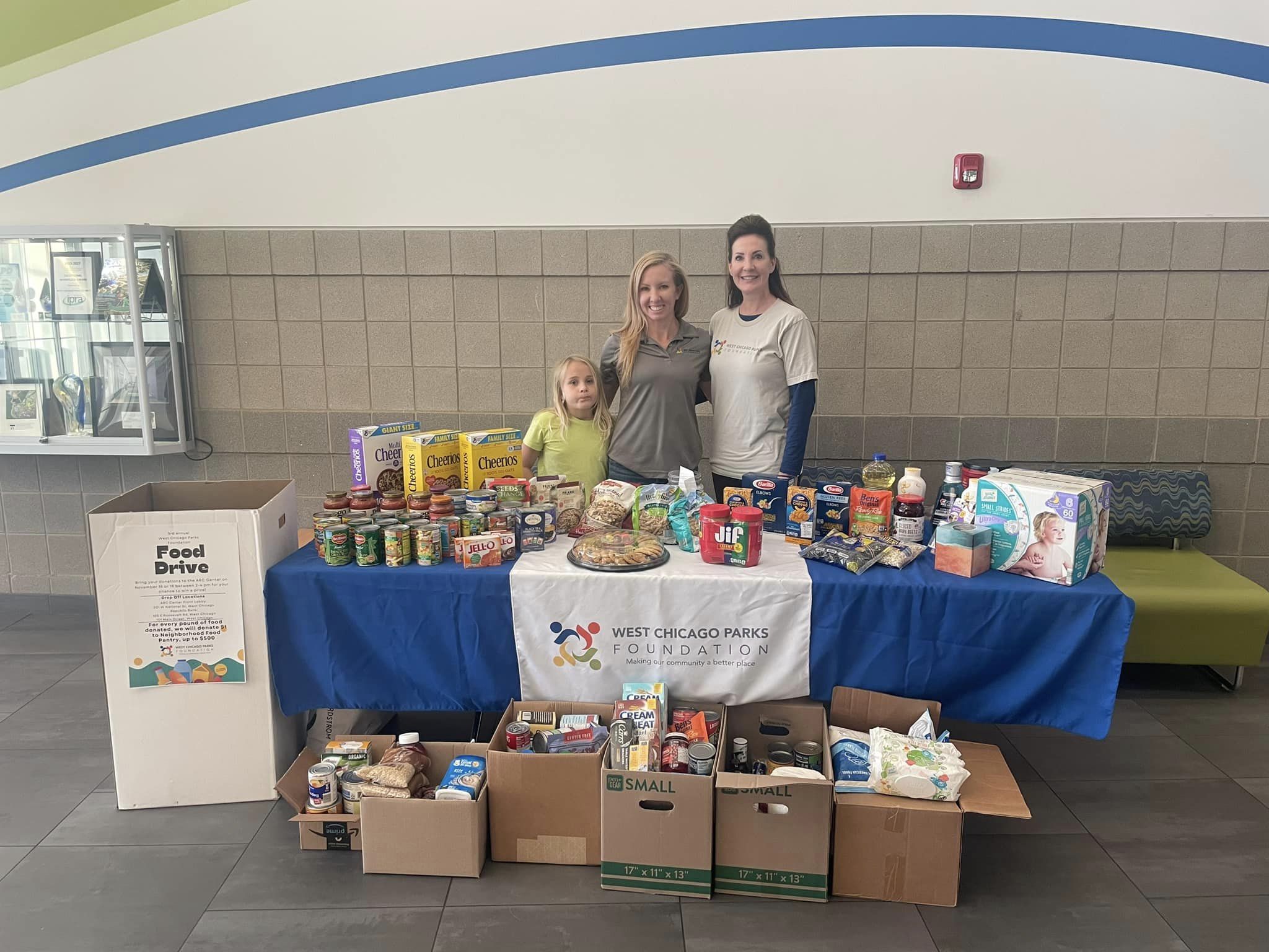 Two Foundation board members and a child stand behind a table filled with food donations.