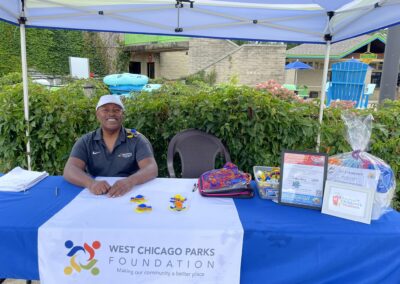 A gentleman is seated, under a canopy, at a table with a Parks Foundation tablecloth. He is smiling posed with some toy turtles on a sunny day. There are prizes to the raight of him.