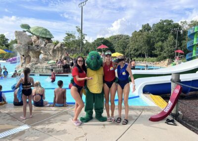 Three lifeguards are posing with a turtle mascot in front of the community waterparks. One lifeguard is holding a toy turtle in their hand.