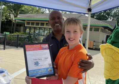 A man is standing behind a boy with a cast on his arm. He is handing him a framed certificate for four passes to Santa's Village Amusement Park.