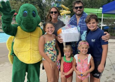 A mascot of a turtle is waving standing next to a family with a mom, dad and four children. They are holding a prize, that is 4 season passes to the community waterpark.